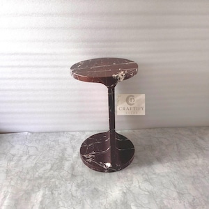 May include: A dark reddish-brown marble side table with white veining, featuring a circular top and a matching circular base connected by a central vertical support. A square label with the text CRAFTIFY DECOR is attached to the central support.