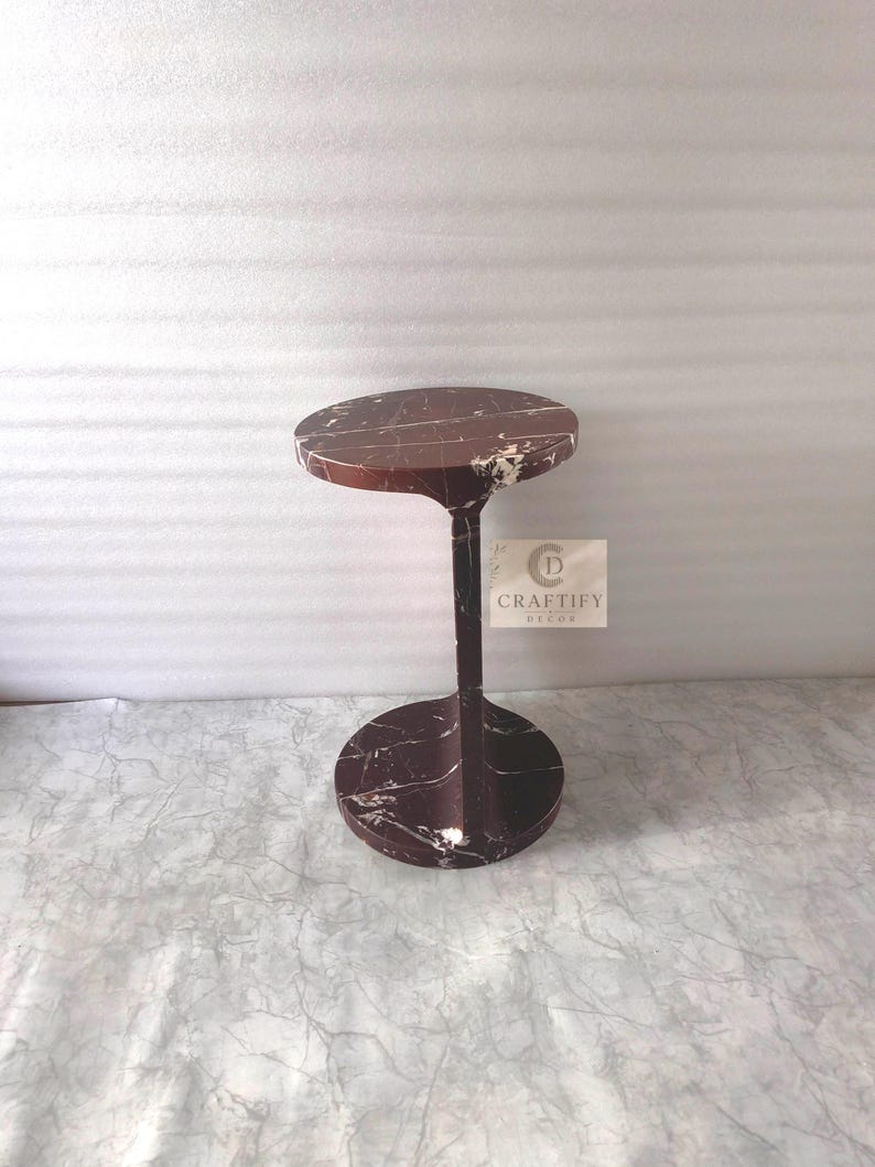 May include: A dark reddish-brown marble side table with white veining, featuring a circular top and a matching circular base connected by a central vertical support. A square label with the text CRAFTIFY DECOR is attached to the central support.