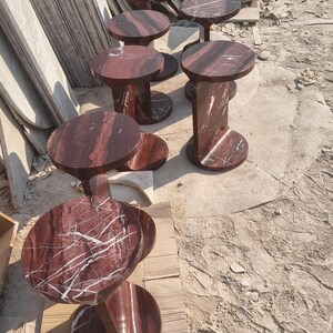May include: A collection of circular side tables crafted from deep red marble with prominent white veining. Each table features a smooth, round top supported by a vertical, curved base that connects to a matching circular floor plate for stability.