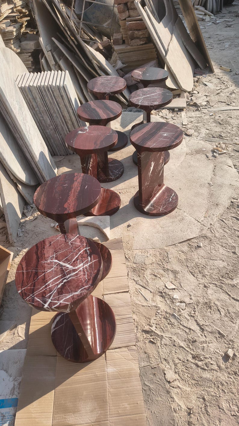 May include: A collection of circular side tables crafted from deep red marble with prominent white veining. Each table features a smooth, round top supported by a vertical, curved base that connects to a matching circular floor plate for stability.