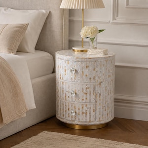 May include: A cylindrical three-drawer nightstand featuring an iridescent mother-of-pearl inlay with a vertical rectangular pattern. The piece includes three clear crystal-style knobs and a circular gold-toned metal base at the bottom.