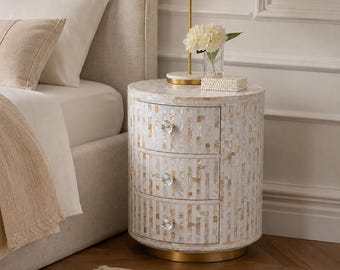 May include: A cylindrical three-drawer nightstand featuring an iridescent mother-of-pearl inlay with a vertical rectangular pattern. The piece includes three clear crystal-style knobs and a circular gold-toned metal base at the bottom.