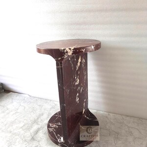 May include: A cylindrical side table crafted from deep red marble with prominent white veining. The piece features a circular top and a matching circular base connected by a vertical central support panel. A small logo reads CRAFTIFY DECOR.
