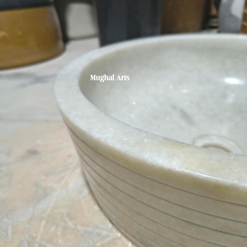 May include: A close-up of a white marble sink with a rounded shape. The sink has a smooth, polished surface and is adorned with horizontal, subtle grooves. The text "Mughal Arts" is visible on the rim.