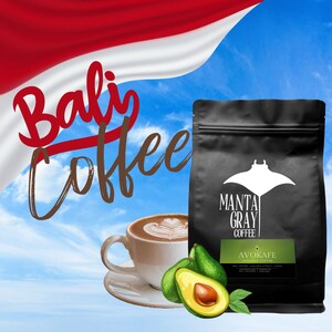 May include: A black bag of Manta Gray Coffee with the text "AVOKAFE" and "BALI COFFEE AVOCADO EXTRACT CREAM" on the front. The bag is in front of a blue sky with white clouds and a red and white flag with the text "Bali Coffee" on it. There is a cup of coffee and two avocados in the foreground.