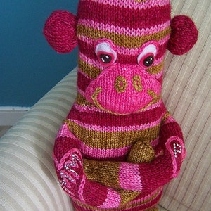 May include: A pink and brown striped knitted monkey toy with a smiling face. The monkey has large pink ears and white eyes with black pupils.