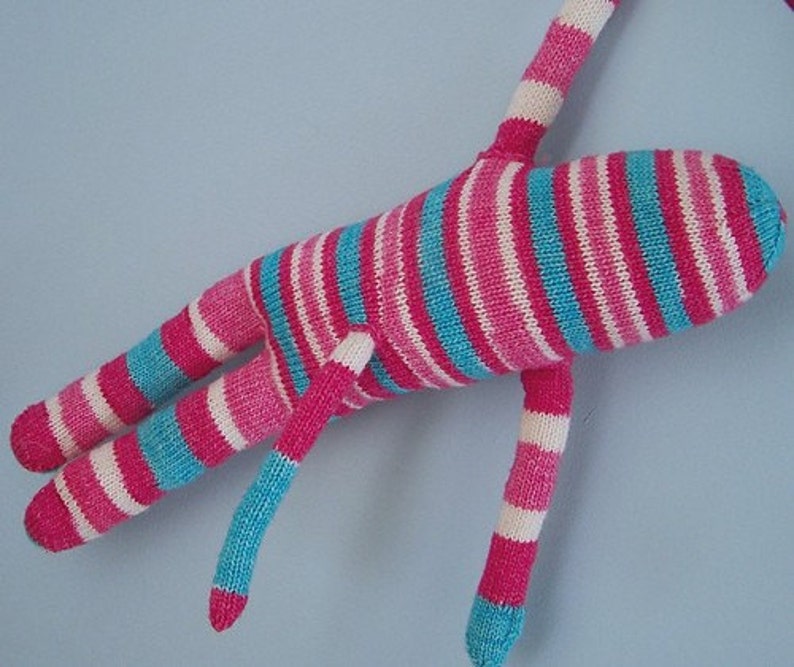 May include: A striped knitted toy with pink, white and blue stripes. The toy has long arms and legs and is lying on a blue surface.