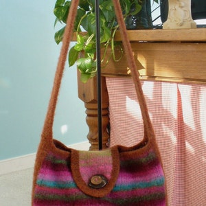 Könnte beinhalten: Eine gestreifte gestrickte Schultertasche mit einer braunen Klappe und einem Holz-Knopfverschluss. Die Tasche hängt an einem schwarzen Ständer.