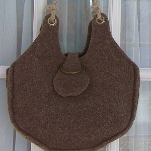 Può includere: Una borsa a mano in feltro marrone con una forma unica e una chiusura a lembo. La borsa ha due anelli in ottone per le maniglie in corda e un anello in ottone sul lembo.