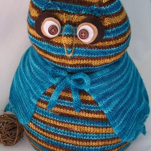 May include: A brown and blue striped knitted owl with a blue knitted cape. The owl has large brown eyes with white button pupils.