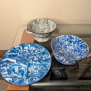 May include: A collection of enamelware kitchen items. Includes a blue and white marbled divided plate, a blue and white speckled bowl with a gold rim, and a green and white speckled colander with a handle and black trim. The items are arranged on a glass surface.