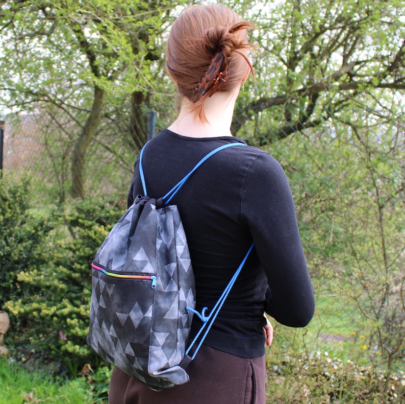 K&ouml;nnte beinhalten: Ein schwarz-grauer Rucksack mit Kordelzug und einem geometrischen Dreiecksmuster. Der Rucksack hat einen Regenbogenrei&szlig;verschluss und blaue Schultergurte. Die Person tr&auml;gt ein schwarzes Langarmshirt und eine braune Hose.