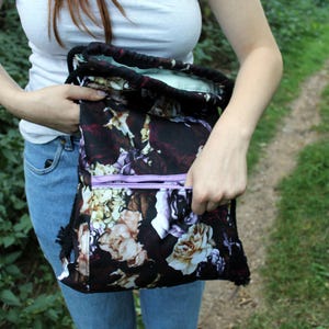 K&ouml;nnte beinhalten: Schwarzer Rucksack mit Kordelzug und floralem Muster in Lila, Creme und Wei&szlig;. Die Tasche hat eine hellviolette Rei&szlig;verschlusstasche und einen schwarzen Kordelzug. Die Innenseite ist hellgr&uuml;n.