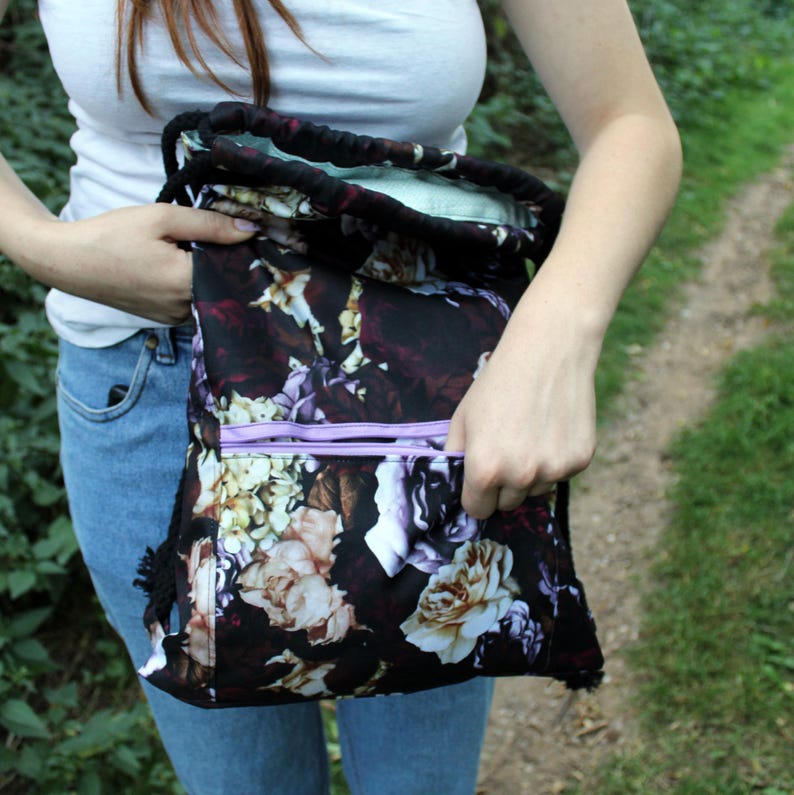 K&ouml;nnte beinhalten: Schwarzer Rucksack mit Kordelzug und floralem Muster in Lila, Creme und Wei&szlig;. Die Tasche hat eine hellviolette Rei&szlig;verschlusstasche und einen schwarzen Kordelzug. Die Innenseite ist hellgr&uuml;n.