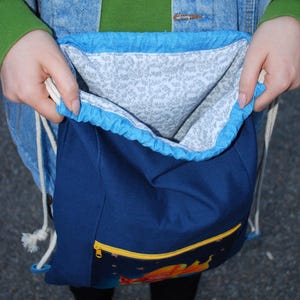 K&ouml;nnte beinhalten: Ein marineblauer Rucksack mit Kordelzug, hellblauer Einfassung und gelber Rei&szlig;verschlusstasche. Das Innenfutter weist ein grau-wei&szlig;es Blumenmuster auf. Die Tasche wird offen gehalten und gibt den Blick ins Innere frei.
