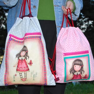 K&ouml;nnte beinhalten: Zwei Rucksackbeutel mit Kordelzug. Einer ist hellblau mit wei&szlig;en Punkten und einem gro&szlig;en Bild eines M&auml;dchens in einem roten Kleid. Der andere ist rosa mit roten Punkten und einem kleineren Bild eines M&auml;dchens in einem roten Kleid. Beide haben rote Kordelz&uuml;ge.