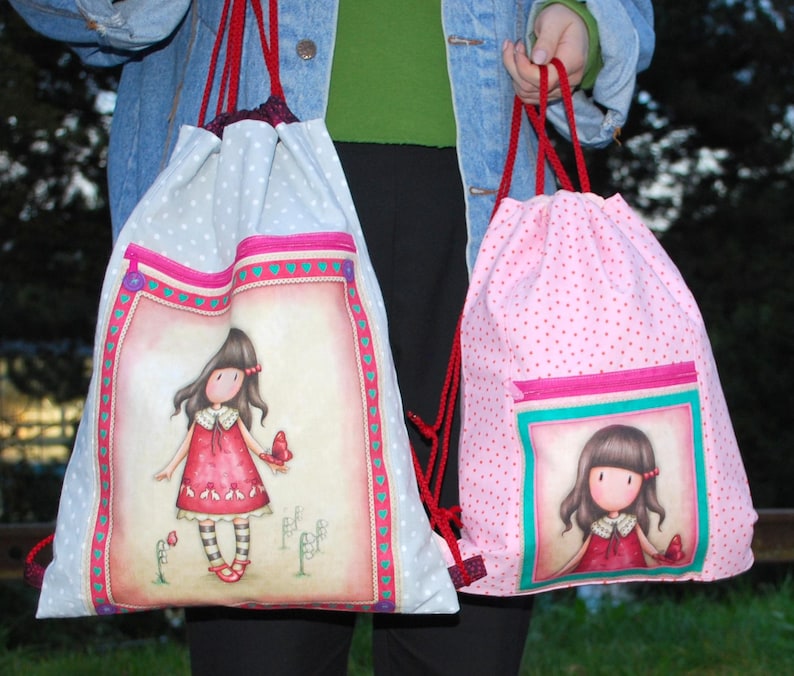 K&ouml;nnte beinhalten: Zwei Rucksackbeutel mit Kordelzug. Einer ist hellblau mit wei&szlig;en Punkten und einem gro&szlig;en Bild eines M&auml;dchens in einem roten Kleid. Der andere ist rosa mit roten Punkten und einem kleineren Bild eines M&auml;dchens in einem roten Kleid. Beide haben rote Kordelz&uuml;ge.