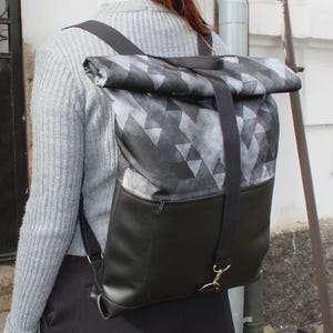 Op de afbeelding: Een zwart-grijze rugzak met een geometrisch patroon op de bovenklep. De onderste helft van de rugzak is effen zwart met een vak met ritssluiting. De rugzak heeft zwarte banden en een goudkleurige sluiting.