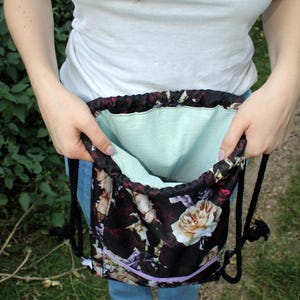 K&ouml;nnte beinhalten: Ein Rucksack mit Kordelzug und floralem Muster mit wei&szlig;en und cremefarbenen Rosen auf dunklem Hintergrund. Die Tasche hat ein hellgr&uuml;nes Innenfutter und einen lila Rei&szlig;verschluss. Schwarze Seilriemen sind sichtbar.