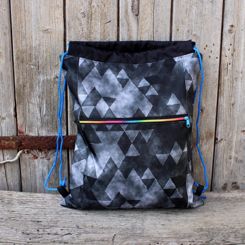 K&ouml;nnte beinhalten: Kordelzug-Rucksack mit geometrischem Muster aus schwarzen und grauen Dreiecken. Die Tasche hat einen schwarzen oberen Teil, eine Vordertasche mit Regenbogenrei&szlig;verschluss und blaue Kordelzugriemen. Die Tasche ist aus Stoff.