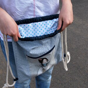 K&ouml;nnte beinhalten: Hellblauer Rucksack mit Kordelzug, wei&szlig;em Herzmuster und einer Rei&szlig;verschlusstasche vorne. Das Innenfutter ist wei&szlig; mit blauen Punkten. Die Tasche hat eine Cartoon-Illustration und wei&szlig;e Seilriemen.