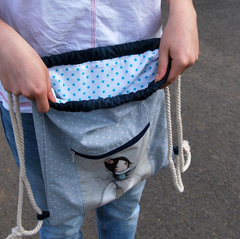 K&ouml;nnte beinhalten: Hellblauer Rucksack mit Kordelzug, wei&szlig;em Herzmuster und einer Rei&szlig;verschlusstasche vorne. Das Innenfutter ist wei&szlig; mit blauen Punkten. Die Tasche hat eine Cartoon-Illustration und wei&szlig;e Seilriemen.