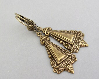 May include: A pair of gold-tone dangle earrings with a triangular design and intricate details. The earrings have a vintage style and are made of metal.