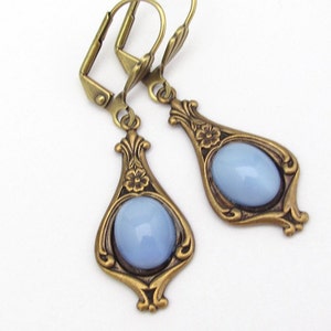 May include: A pair of antique-style earrings featuring light blue oval stones set in ornate, teardrop-shaped bronze frames. Each frame has floral detailing and a lever-back closure.