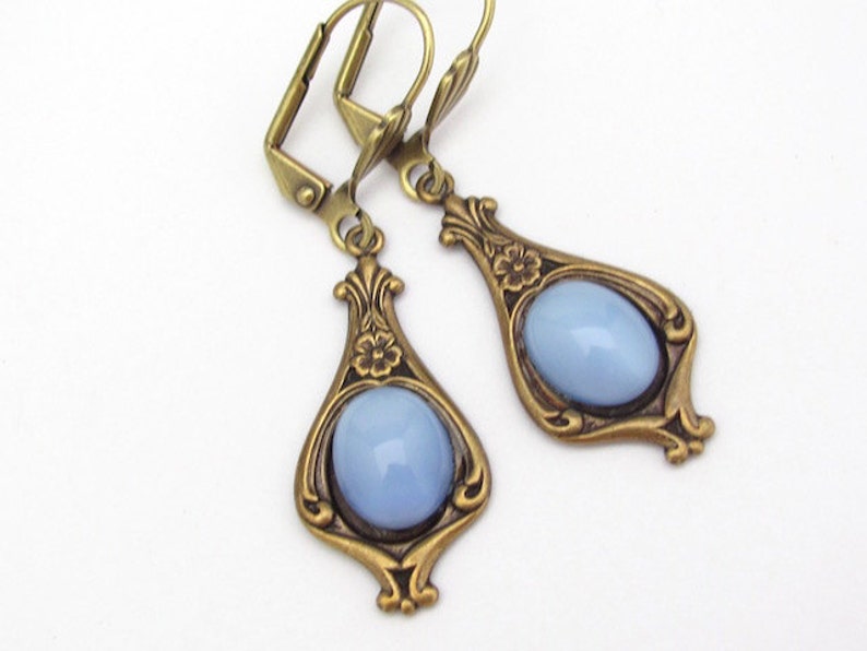 May include: A pair of antique-style earrings featuring light blue oval stones set in ornate, teardrop-shaped bronze frames. Each frame has floral detailing and a lever-back closure.