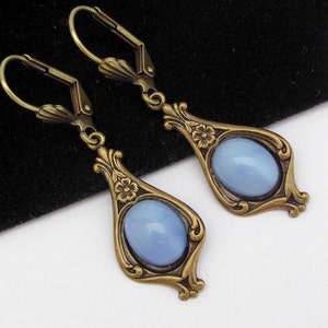 May include: A pair of vintage-style drop earrings. Each earring has a pale blue oval stone set in an ornate, bronze-coloured metal frame with floral details. The earrings have a lever-back clasp.