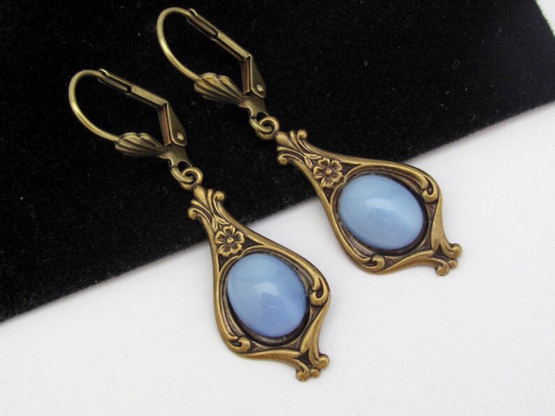 May include: A pair of vintage-style drop earrings. Each earring has a pale blue oval stone set in an ornate, bronze-coloured metal frame with floral details. The earrings have a lever-back clasp.