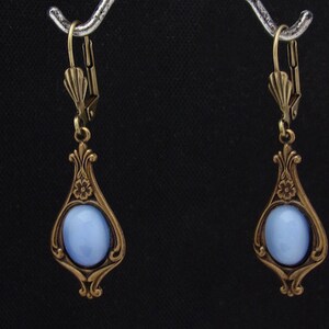 May include: A pair of vintage-style drop earrings. Each earring has a pale blue oval stone set in an ornate, teardrop-shaped bronze frame. The frames feature floral and scroll detailing. The earrings have lever-back closures.