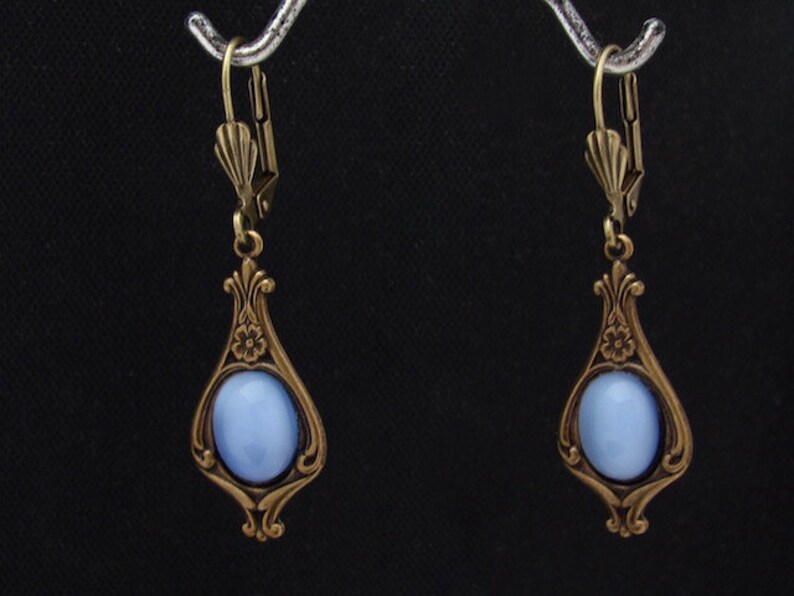 May include: A pair of vintage-style drop earrings. Each earring has a pale blue oval stone set in an ornate, teardrop-shaped bronze frame. The frames feature floral and scroll detailing. The earrings have lever-back closures.
