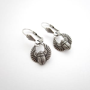 May include: A pair of silver-toned metal earrings featuring a scarab beetle design with outstretched wings. The earrings are suspended from lever-back closures, showcasing a detailed, textured finish on the beetle bodies and wings.