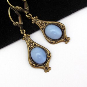 May include: A pair of vintage-style drop earrings. Each earring has a pale blue oval stone set in an ornate, bronze-coloured frame. The frames feature floral details and decorative flourishes.