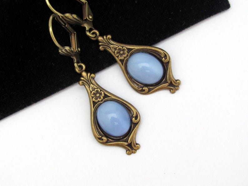 May include: A pair of vintage-style drop earrings. Each earring has a pale blue oval stone set in an ornate, bronze-coloured frame. The frames feature floral details and decorative flourishes.