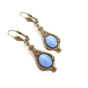 May include: A pair of vintage-style earrings featuring a pale blue oval stone set within an ornate, bronze-toned frame. The earrings have a lever-back clasp and floral detailing. The design is symmetrical and elegant, suitable for a vintage aesthetic.