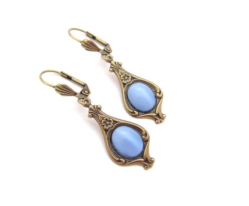 May include: A pair of vintage-style earrings featuring a pale blue oval stone set within an ornate, bronze-toned frame. The earrings have a lever-back clasp and floral detailing. The design is symmetrical and elegant, suitable for a vintage aesthetic.