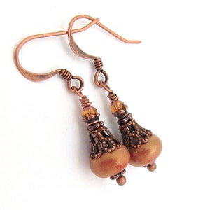 May include: A pair of handcrafted copper earrings. Each earring features a teardrop-shaped, light brown bead with intricate filigree detailing and a small amber-colored crystal. The earrings have copper ear wires and are designed for pierced ears.