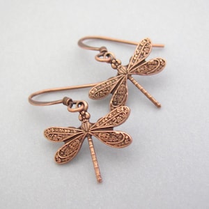 May include: A pair of copper-colored dragonfly earrings. Each earring features a detailed dragonfly design with intricate wing patterns and a hook closure. The earrings are displayed against a light gray background.