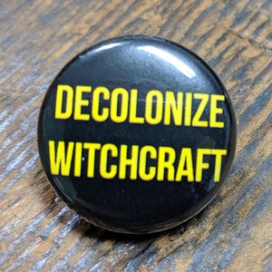 Puede incluir: Un botón negro con el texto "DECOLONIZE WITCHCRAFT" en letras amarillas.