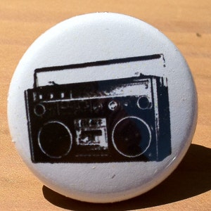 May include: A white button with a black and white image of a vintage boombox. The boombox has two speakers, a cassette player, and a radio dial.