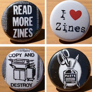 May include: Four black and white button badges with different designs. The first badge says "Read More Zines". The second badge says "I ♥ Zines". The third badge says "Copy and Destroy" and has a picture of a photocopier. The fourth badge has a picture of scissors, a pencil, and a bottle of glue.