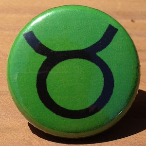 Puede incluir: Un botón verde con un símbolo negro del zodíaco de Tauro.