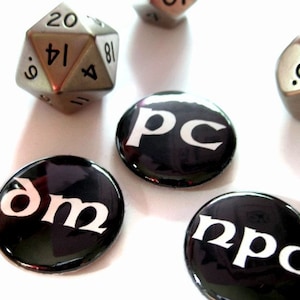 Puede incluir: Cuatro botones negros y blancos con las palabras "DM", "PC", "NPC" y "GM" impresas en blanco sobre un fondo negro. Los botones están sobre una superficie blanca con un dado de 20 caras plateado.