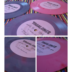 Puede incluir: Cuatro discos de vinilo rosa y morado con etiquetas blancas. Las etiquetas tienen el texto "Tagalongs" y "Side B 33 1/3 rpm" escrito en ellas. Los discos están apilados uno encima del otro.
