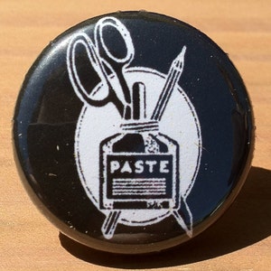 May include: Black and white button with a graphic of a bottle of paste, scissors, and a pencil. The bottle has the word "PASTE" printed on it.