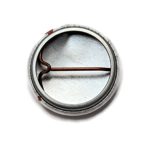 May include: The back of a silver button with a copper pin. The button has a smooth, shiny surface.