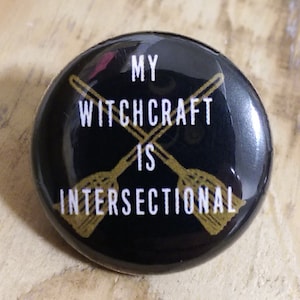 May include: Black button with a gold design of two crossed broomsticks and the text "My Witchcraft is Intersectional".