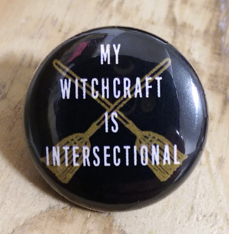 Puede incluir: Bot&oacute;n negro con un dise&ntilde;o dorado de dos escobas cruzadas y el texto "My Witchcraft is Intersectional".